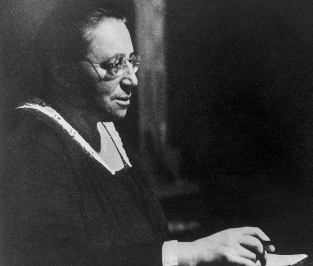 Black and white photo of a woman with glasses in a black cloak