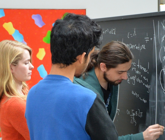 Three men and a woman standing at a chalkboard working on equations