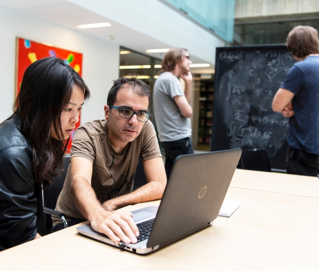 Man and woman working at a computer with two men working at a blackboard in the background