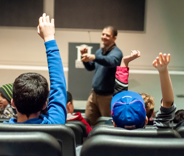 Kids in a lecture hall, raising their hands, asking questions