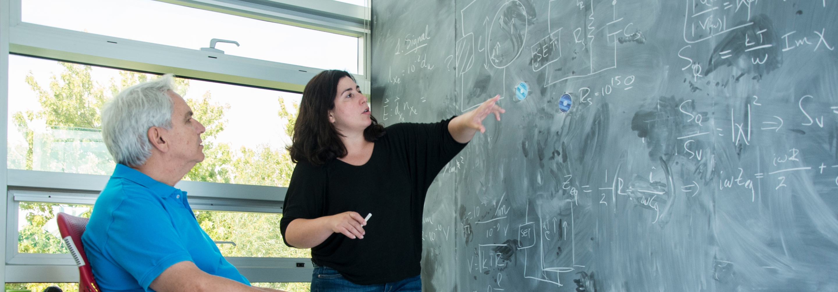 Woman standing at a blackboard pointing at equations, man sitting on chair listening