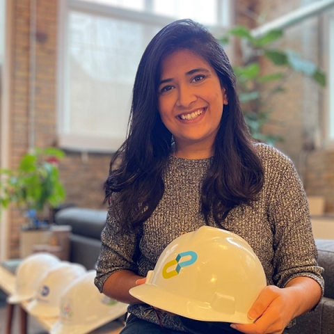 Woman in a grey knit sweater holding a hard hat and standing in an office