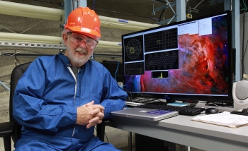 Arthur B. McDonald with a hard hat on, sitting in front of his computer