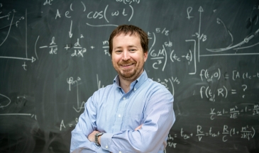 Man in a light blue dress shirt standing in front of a chalkboard with equations with his arms crossed